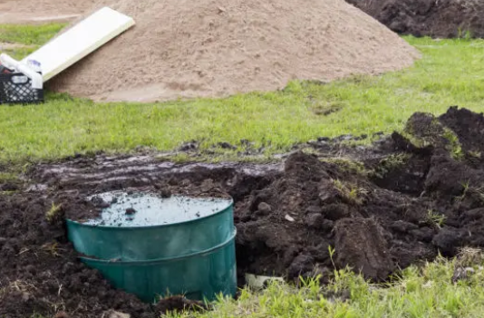 New mound septic system under construction with a green tank access riser, excavated soil, and a mound of sand in a yard