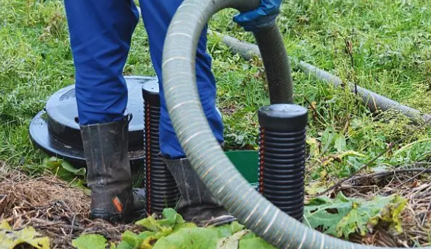 Technician in blue gloves and rubber boots feeding a suction hose into septic drain field risers with black corrugated pipes